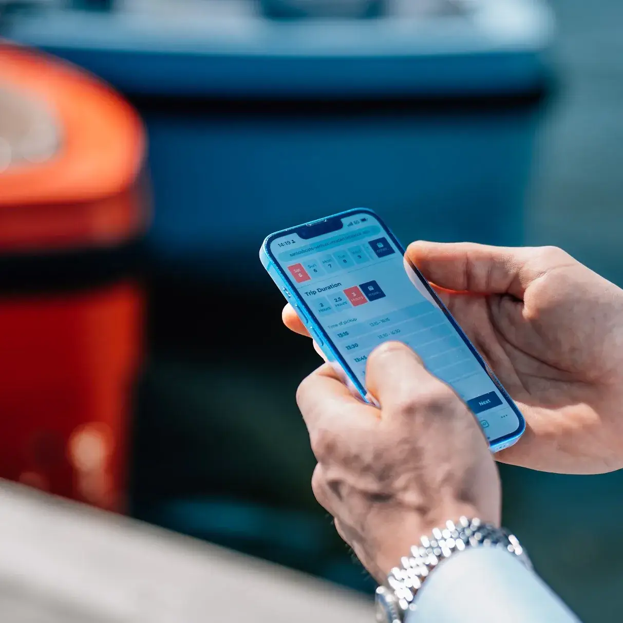 A hand holds a phone with a boat booking open, orange rental boats behind