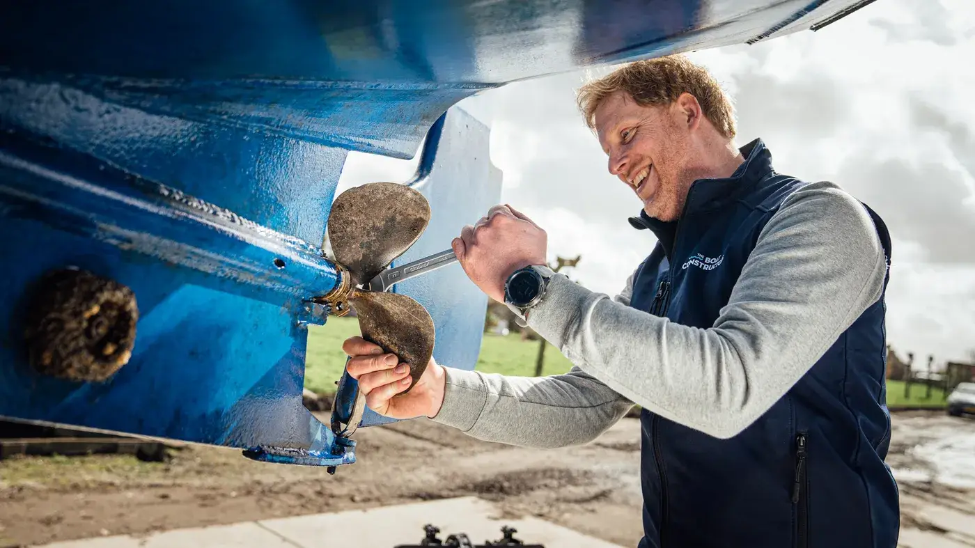 Een lachende monteur werkt aan de schroef onder de romp van een boot op de kant