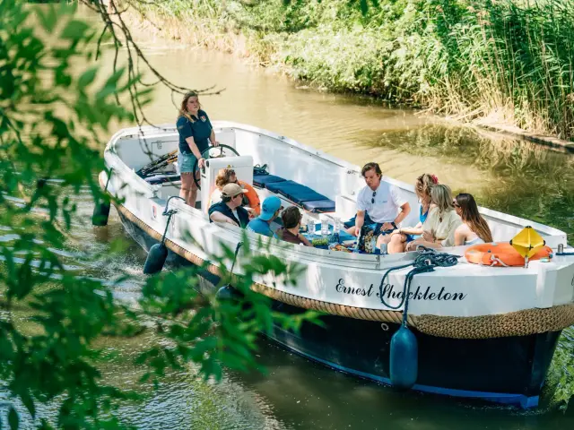 An open tour boat on a green river, a skipper at the wheel and a group of guests aboard, seen through the leaves